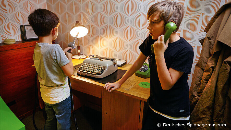 Zwei Kinder im Deutschen Spionagemuseum: Sie versuchen in einem DDR-Büro durch Untersuchen und Telefonieren ein Rätsel zu lösen.