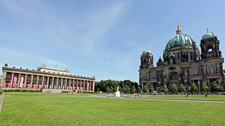 Berlin: Links das Alte Museum, rechts das Schloss Charlottenburg. Der Himmel ist blau und die Wiese grün.