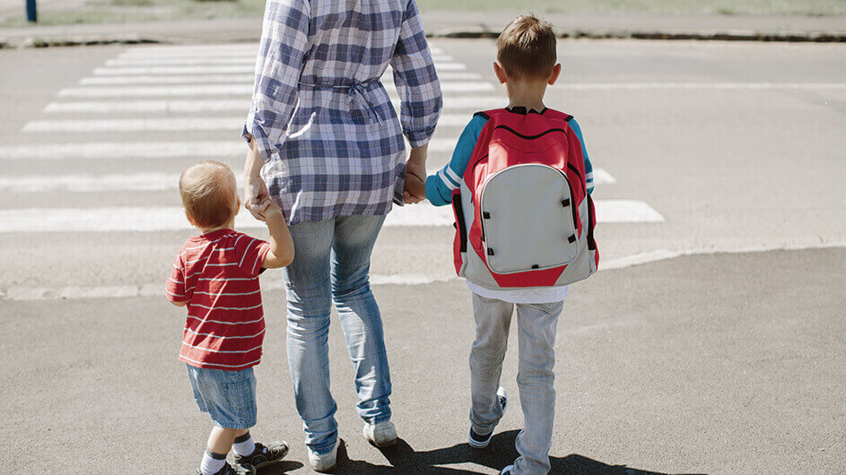 eine Mutter geht mit ihren zwei kleinen Söhnen, die Schulranzen tragen, an der Hand über einen Zebrastreifen