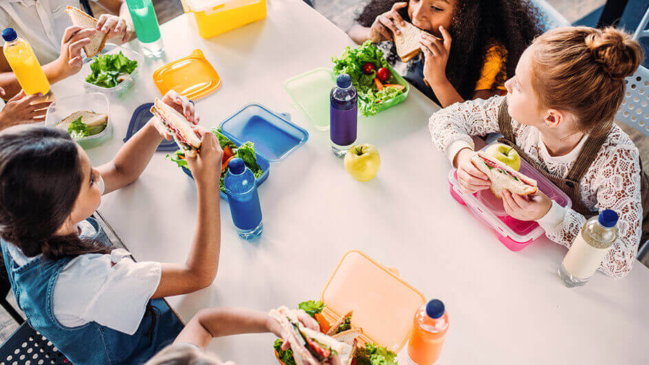 Aus der Vogelperspektive: Kinder sitzen nebeneinander am Tisch uns essen aus ihren Lunchboxen ihr Mittagessen in der Schule. Daneben stehen Flaschen mit Getränken.