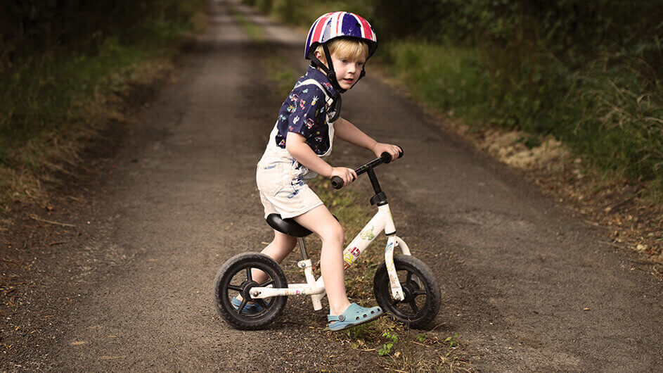 Ein kleiner Junge steht über seinem Laufrad. Er trägt einen Helm. Er ist auf einem Waldweg und wartet auf seine Eltern.