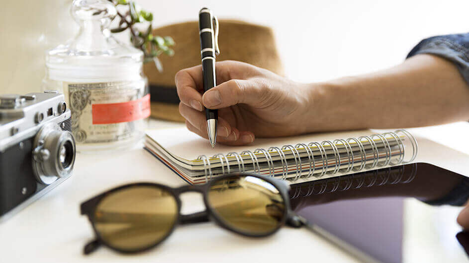 Ein am Schreibtisch sitzender Mann plant eine Stadtrallye. Er schreibt auf einen Block. Auf dem Schreibtisch liegen außerdem ein Hut, eine Sonnenbrille, ein Tablett und eine Kamera.