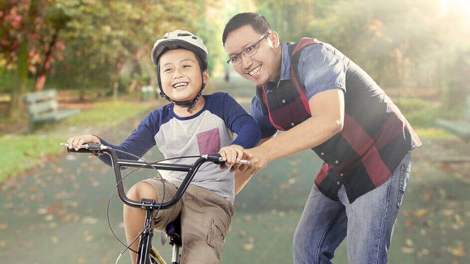 Im Park: Ein Vater schiebt seinen Sohn, der auf einem Fahrrad sitzt, an. Beide lächeln.