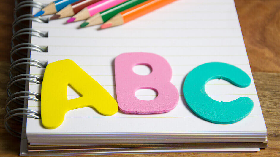 Aus Moosgummi ausgeschnittene, bunte Buchstaben (A, B und C) auf einem Schulblock. Dahinter liegen Buntstifte.