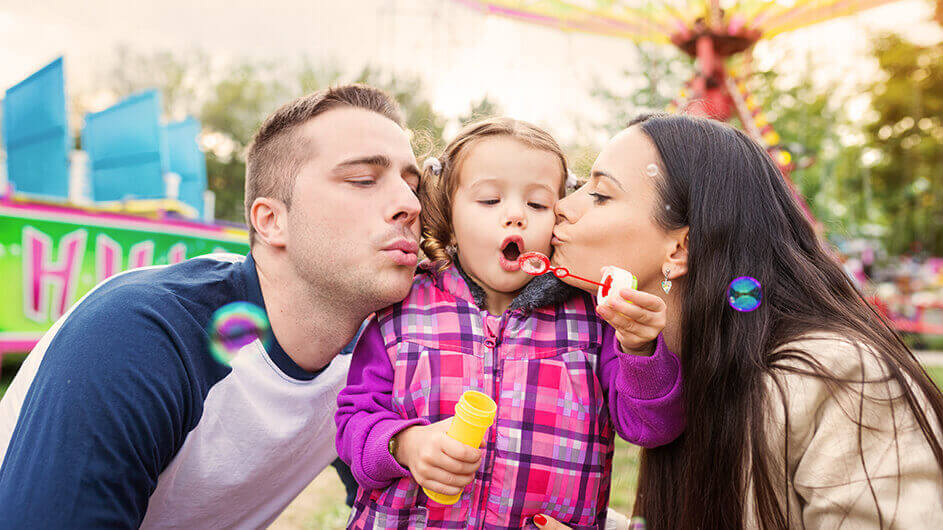Ein Kleinkind bläst Seifenblasen in einem Freizeitpark. Links steht die Mutter und gibt dem Kind ein Küsschen. Rechts steht der Vater und pustet mit.