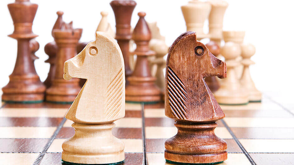 Zwei Springer-Schachfiguren (weiß und schwarz) stehen nebeneinander auf einem Schachbrett. Im Hintergrund sind andere Schachfiguren zu sehen.