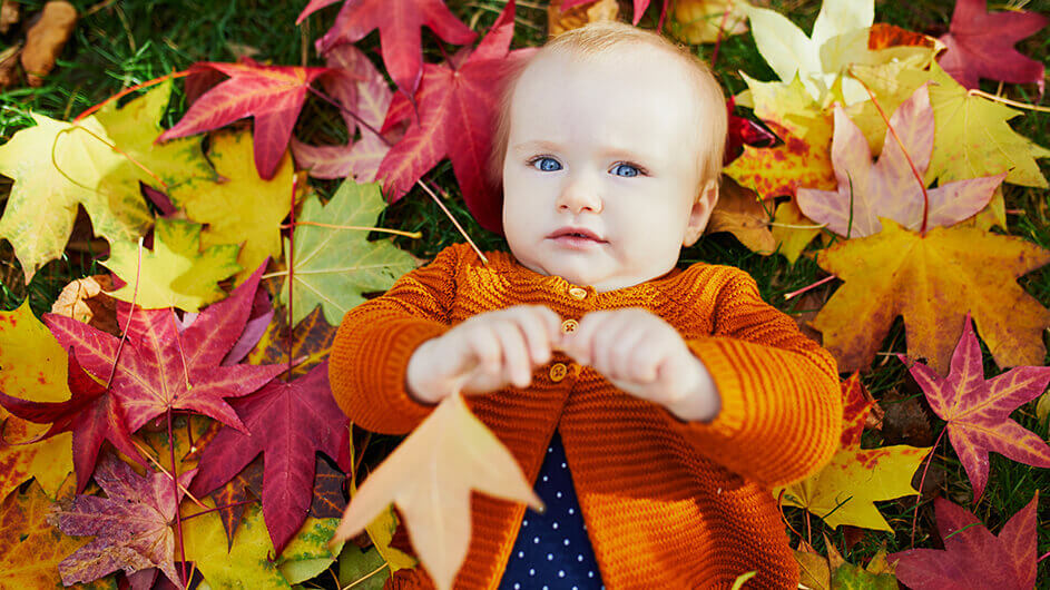 Baby auf Herbstwiese liegend mit buntem Herbstlaub: Es trägt ein gepunktetes Oberteil und eine orangefarbene Strickjacke darüber.