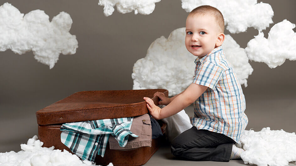 Ein Junge packt einen Koffer fürs Reisen - um ihn herum ist Dekoration in Wolkenform. Er schaut in die Kamera und lächelt fröhlich.