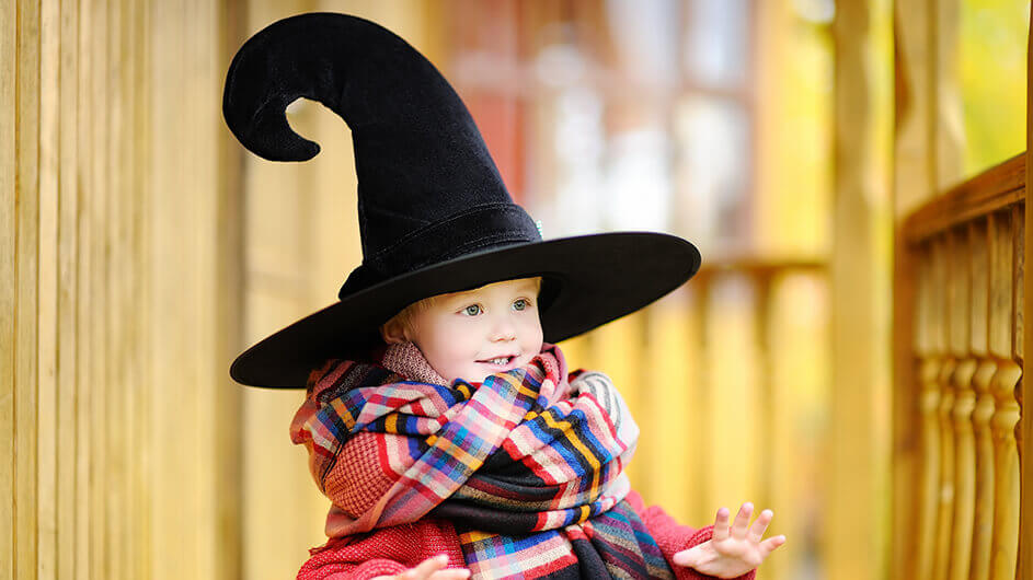 Ein kleiner Junge, der sich für Halloween als Zauberer verkleidet hat. Er trägt einen bunten Schal und einen riesigen Zauber-Hut mit gebogenem Zipfel.