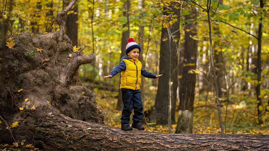 Ein kleiner Junge mit knallgelber Weste und Mütze mit roter Bommel balanciert im Herbstwald auf einem Baumstamm.