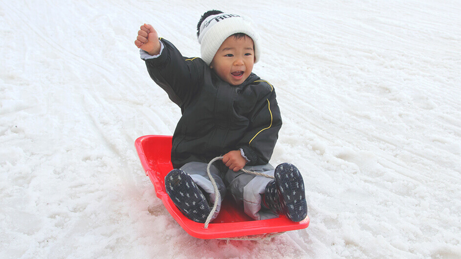 Japanischer kleiner Junge auf einem roten Schlitten im Winter: Das Kind hebt freudig einen Arm hoch und jauchzt.