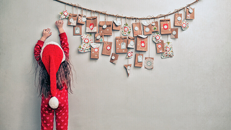 Kind streckt sich nach selbstgemachtem Adventskalender: Das Mädchen trägt eine Weihnachtsmütze. Der kalender besteht aus einem ast, an dem bunte Tüten mit Überraschungen hängen.