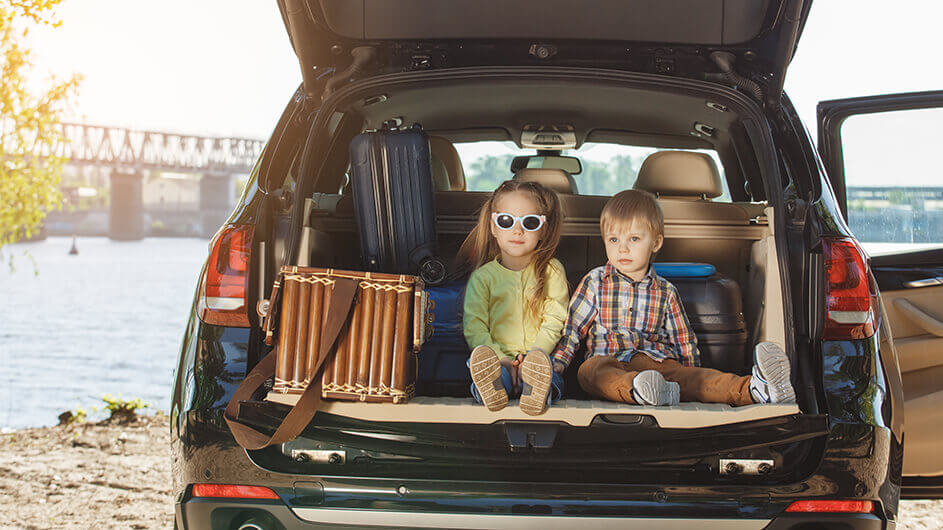 Zwei Kinder sitzen bei der Anreise in den Familienurlaub im offenen Kofferraum des Familienautos. Neben ihnen liegen Koffer. Das Mädchen trägt eine Sonnenbrille, der Junge sieht verträumt aus. Das Auto steht vor einem Fluss. Die Sonne scheint.