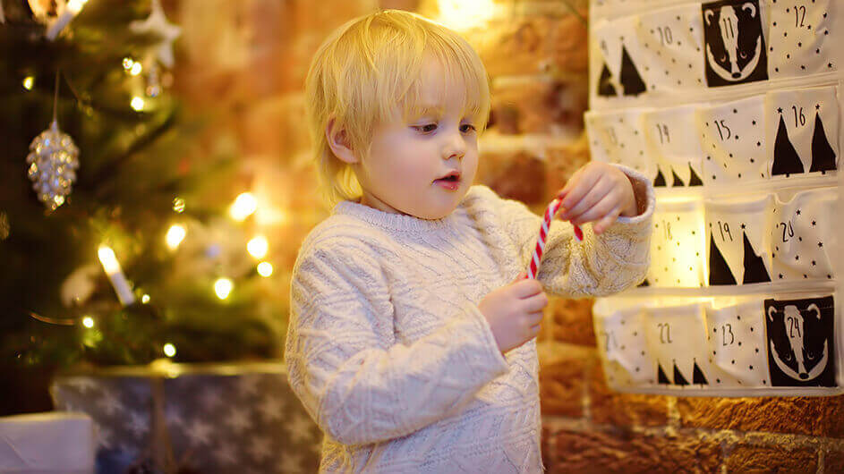 Ein kleines Kind betrachtet eine Zuckerstange, die es aus einem Adventskalender geholt hat. Hinter ihm steht ein Weihnachtsbaum.