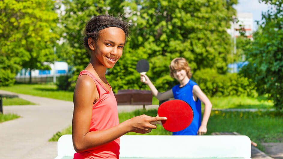 Ein Mädchen und ein Junge draußen beim Tischtennisspielen. Beide schauen in die Kamera und lächeln.
