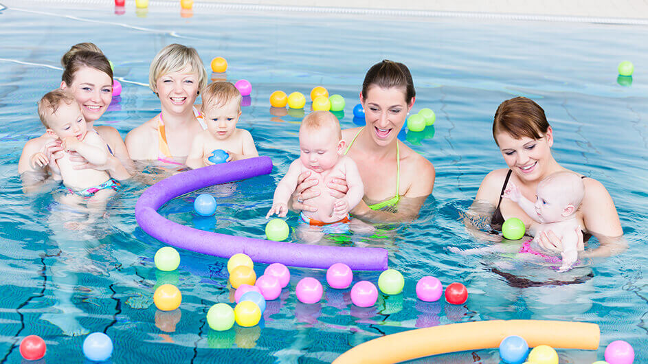 Vier Mütter mit ihren kleinen Kindern beim Babyschwimmen im Schwimmbad. Die Mütter lächeln und die Babys planschen und spielen mit buntem Wasserspielzeug (Bälle und Schwimmnudeln).