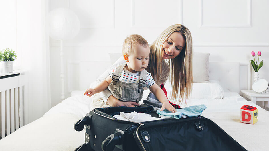 Mutter und kleines Kind packen gemeinsam den Koffer für eine Reise: Die Mutter lächelt und das Kind guckt konzentriert. Sie sitzen auf einem Bett in einem hellen, freundlichen Raum.