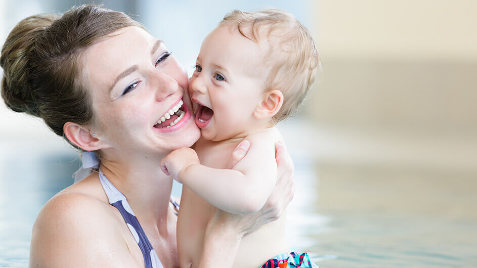 Eine Mutter mit ihrem Baby im Schwimmbad beim Babyschwimmen. Die Mutter hält das Kind lächelnd in den Armen, das sich an ihr Gesicht schmiegt. Das Kind lacht dabei.