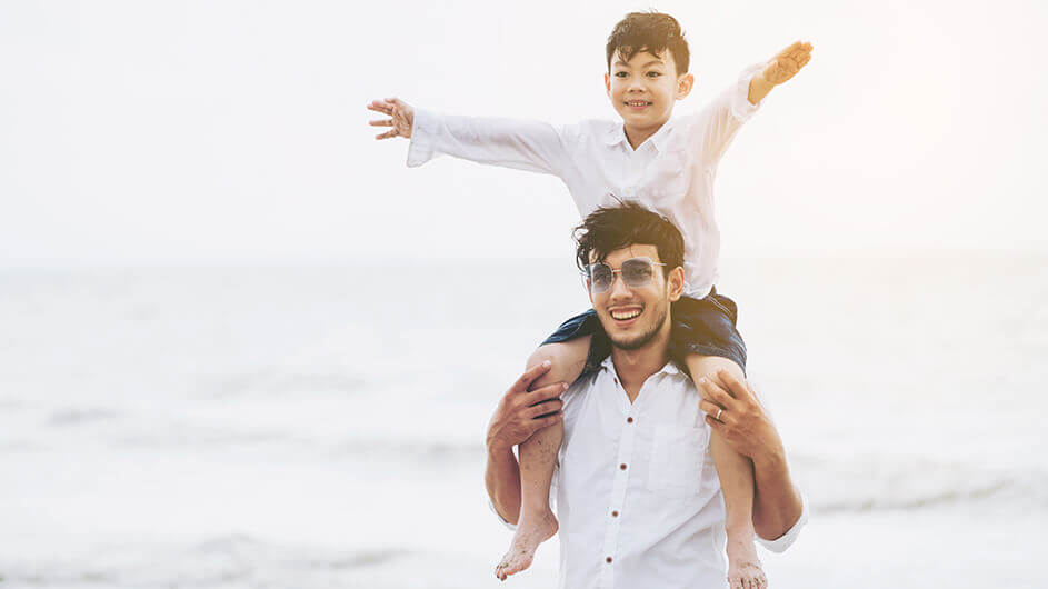 Der kleine Sohn sitzt auf den Schultern seines Vaters am Strand vorm Meer. Beide lächeln und das Kind streckt seine Arme in die Luft.