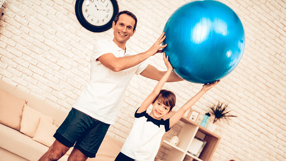 Vater und Sohn Zuhause mit einem Gymnastikball: Der sohn hält den großen, blauen Ball hoch und der Vater hält ihn zusätzlich fest. Beide stehen in einem schicken, hellen Wohnzimmer.