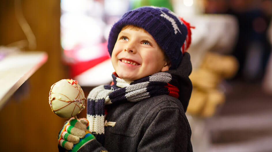 Ein kleiner, lächelnder junge mit einer Schokofrucht auf einem Weihnachtsmarkt bzw. Wintermarkt. Er trägt Mütze, Schal und Handschuhe