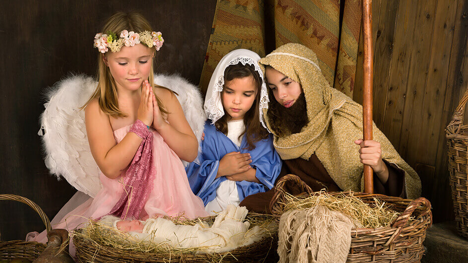 Kinder als Engel, Maria und Josef beim weihnachtlichen Krippenspiel: Der engel betet und schaut auf das Jesuskind. Maria und Josef betrachten ebenfalls ihr Kind.
