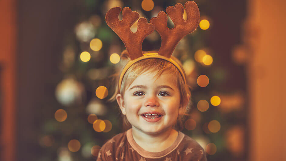 Ei n lachendes Kleinkind mit Rentier-Geweih-Haarreif und rot geschminkter Nase. Im Hintergrund glitzern Weihnachtslichter.