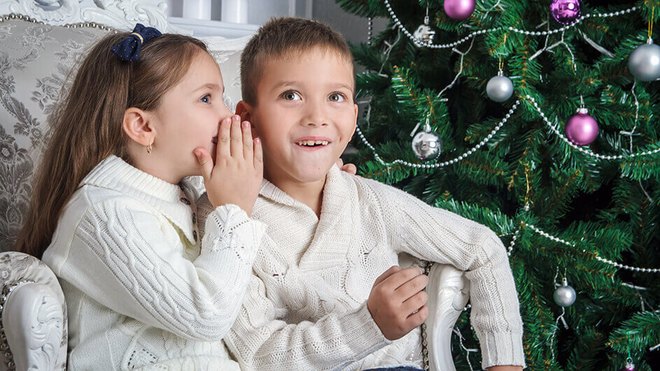 Die Schwester flüstert ihrem bruder etwas ins Ohr. Beide lächeln und sitzen vorm geschmückten Weihnachtsbaum.