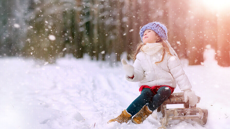 Ein kleines Mädcehn sitzt auf einem Schlitten und schaut siczh verträumt die fallenden Schneeflocken an. Die Sonne scheint und sie trägt eine hellblaue Mütze und Handschuhe.
