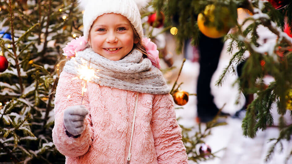 Ein lächelndes Mädchen mit einer Wunderkerze auf einem Wintermarkt bzw. Weihnachtsmarkt. Es steht neben einem geschmückten Weihnachtsbaum