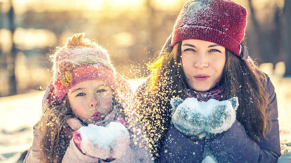 Mutter und Tochter pusten Schneeflocken Richtung Kamera: Beide tragen Mützen. Die Sonne scheint.