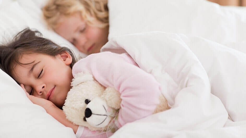 Zwei schlafende Geschwister, die nebeneinander liegen. Das vordere Kind lächelt und hält einen Teddybären im Arm.