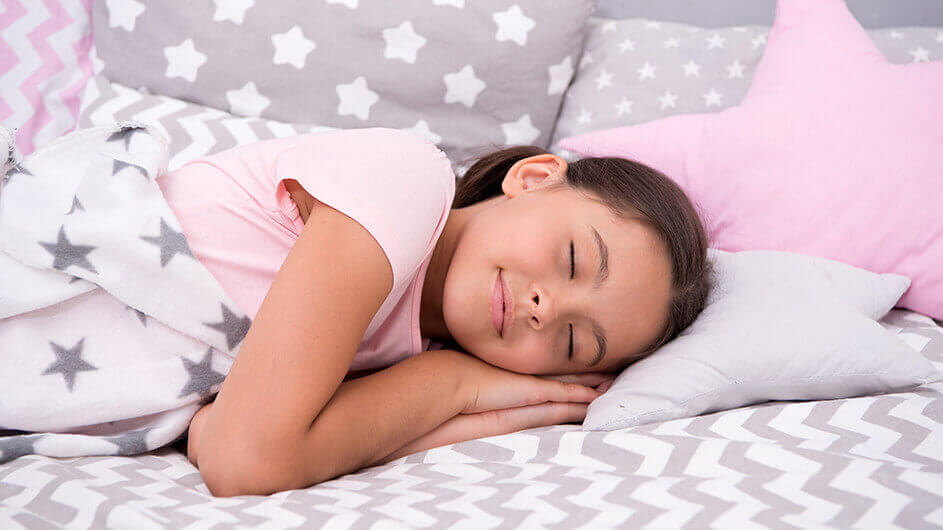 7 Einschlaftipps: Ein schlafendes Mädchen: Das Kind lächelt im Schlaf und liegt in einem bett mit vielen Kissen. Verziert sind diese mit Sternen.