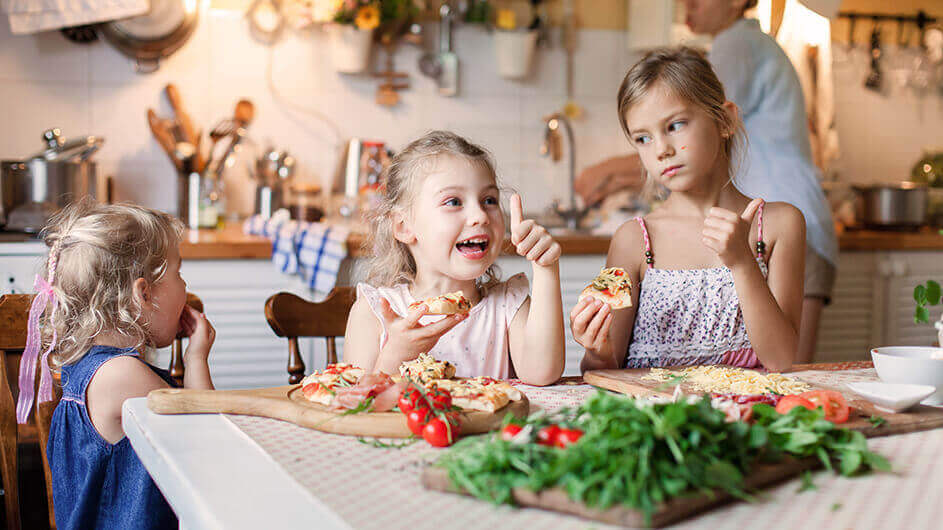 Drei Mädchen beim Kochen bzw. Backen von Pizza: Das Mittlere Kind zeigt lachend den Daumen nach oben.