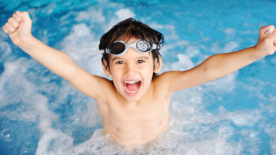 Ein glücklich jubelnder Junge mit Schwimmbrille im Schwimmbad: Er ist im Wasser und streckt die Arme fröhlich nach oben.