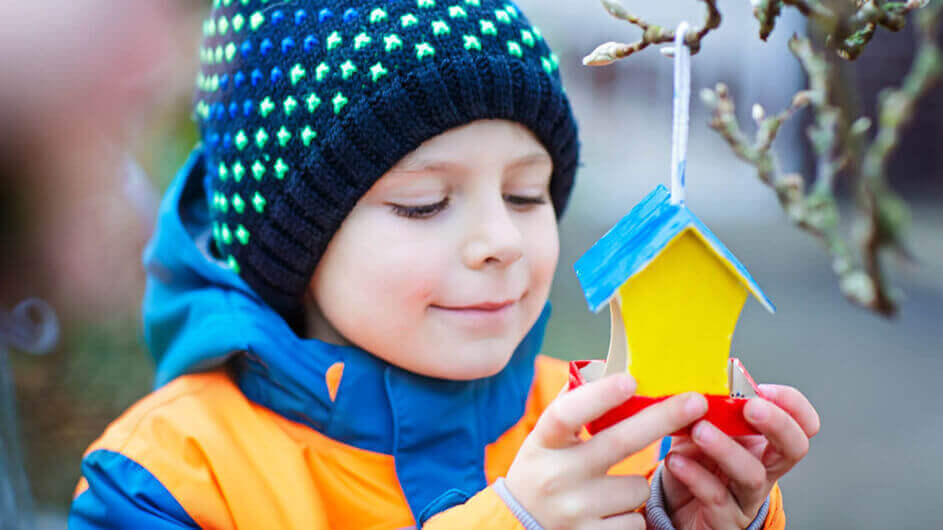Ein Junge hält im Winter lächelnd ein Vogelhaus in seinen Händen, das an einem Zweig hängt
