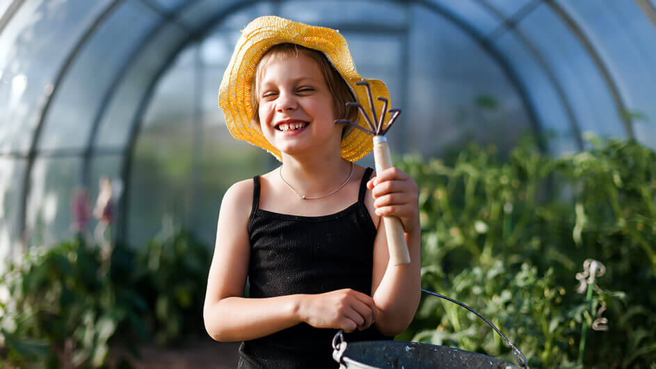 Ein fröhlich lachendes Mädchen bei der Gartenarbeit: Es trägt einen Strohhut und hält eine Harke sowie einen Eimer