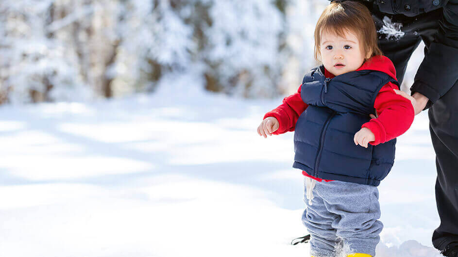 Ein Kleinkind steht im Schnee. Der Vater hält es am Arm fest.