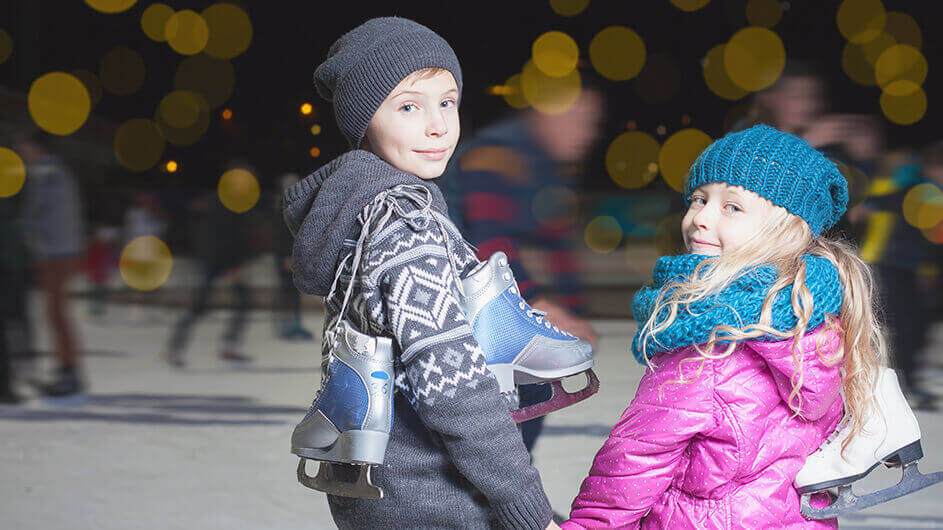 Zwei Kinder halten Händchen vor der Eisbahn und tragen Schlittschuhe mit sich: Beide gucken in die Kamera und lächeln.