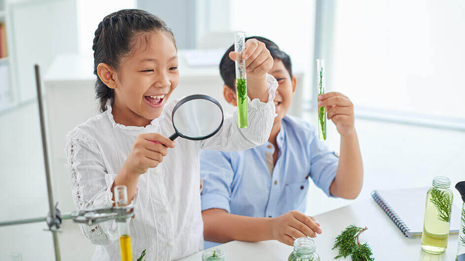 Kinder beim Experimentieren in einer Kinder-universität mit Reagenzgläsern und einer Lupe. Sie freuen sich.