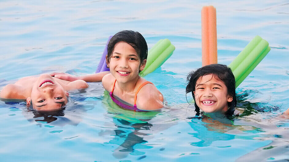 Drei fröhliche Kinder beim Schwimmen im Schwimmbad mit bunten Schwimmhilfen: Sie lächeln. Ein Kind schwimmt kopfüber.