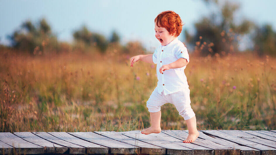 Ein kleiner Junge läuft barfuß jubelnd über einen Steg durch eine Sommerwiese
