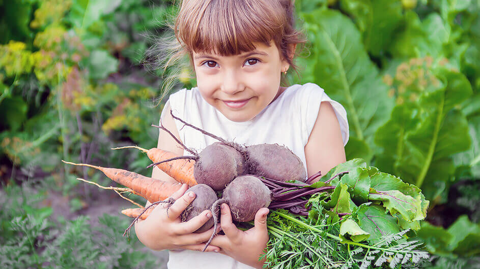 Ein glücklich lächelndes Mädchen steht mit Karotten und Roter Beete im Arm vorm Gemüsegarten