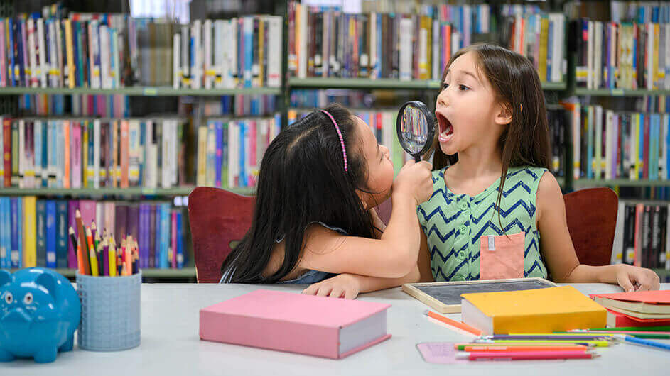 Zwei Mädchen lesen und spielen in einer Bibliothek mit einer Lupe
