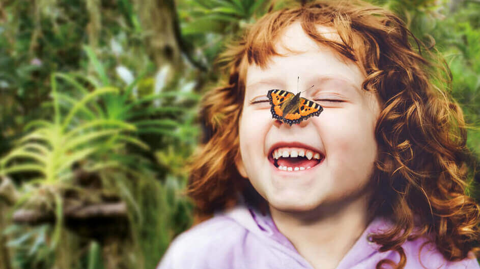Lachendes Mädchen mit einem Schmetterling auf seiner Nase: Der Schmetterling ist ein Kleiner Fuchs und das Kind hat rote, lockige Haare und steht in einem Tropenhaus.