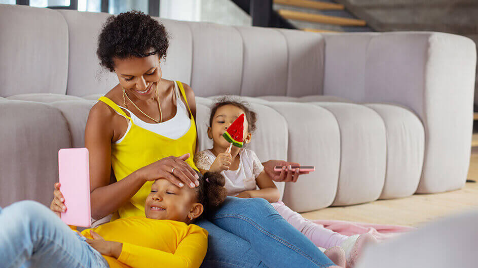 Mutter und zwei Töchter beim Spielen von Kinder-Apps: Alle lächeln und liegen entspannt auf dem Sofa.