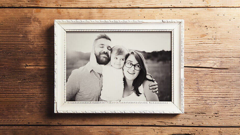 Ein schwarz-weißes Familienfoto in einem Bilderrahmen auf Holzuntergrund liegend: Die Familie aus Vater, Mutter und Kleinkind sieht glücklich aus.
