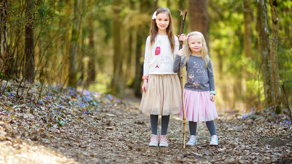 Zwei Mädchen stehen lächelnd mit einem Stock im Wald