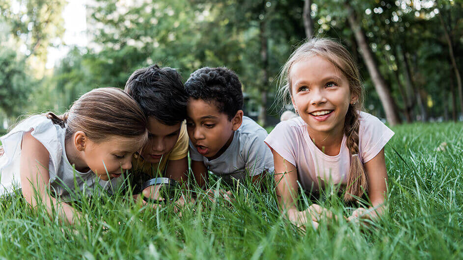 Kinder liegen im Gras und beobachten staunend und lächelnd mit einer Lupe Insekten