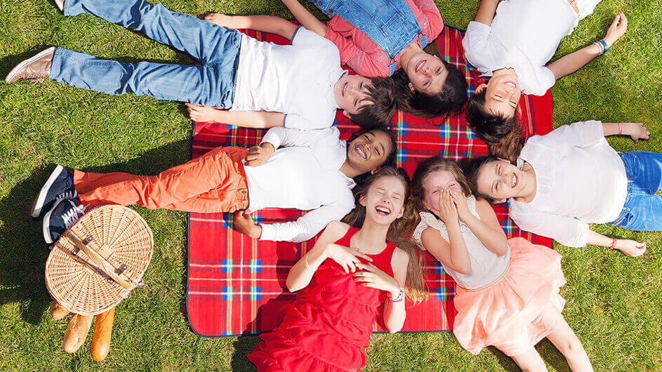 Lachende Kinder liegen beim Picknicken im Kreis auf einer Picknickdecke neben einem Picknickkorb
